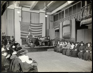 Folk i et auditorium i New York Association for the Blind med juletræ og folk, der muligvis opfører et sketch på scenen med orgel, 1920 (sølv gelatinetryk)